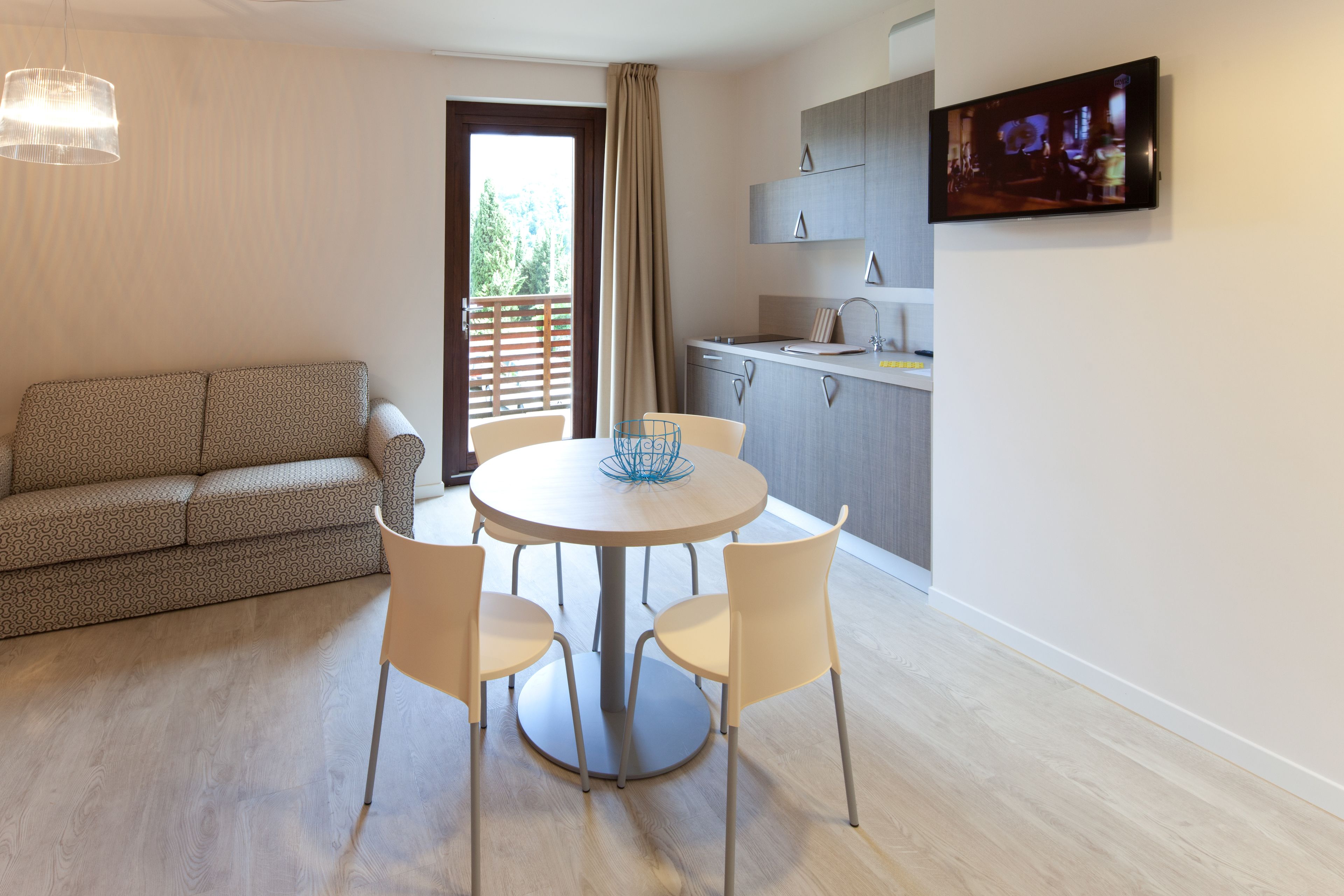 A modern living room with a sofa, a round dining table with chairs, a kitchen area with cabinets and a TV, and a balcony with a curtain.