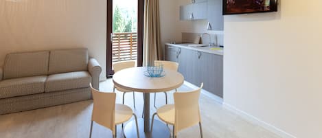 A modern living room with a sofa, a round dining table with chairs, a kitchen area with cabinets and a TV, and a balcony with a curtain.
