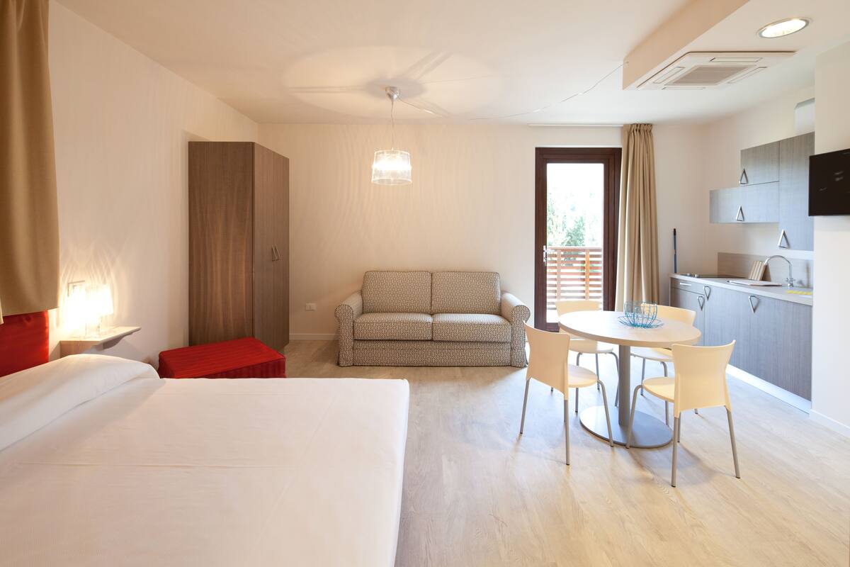 A hotel room with a bed, a sofa, a dining table with chairs, a kitchen area, and a ceiling fan.