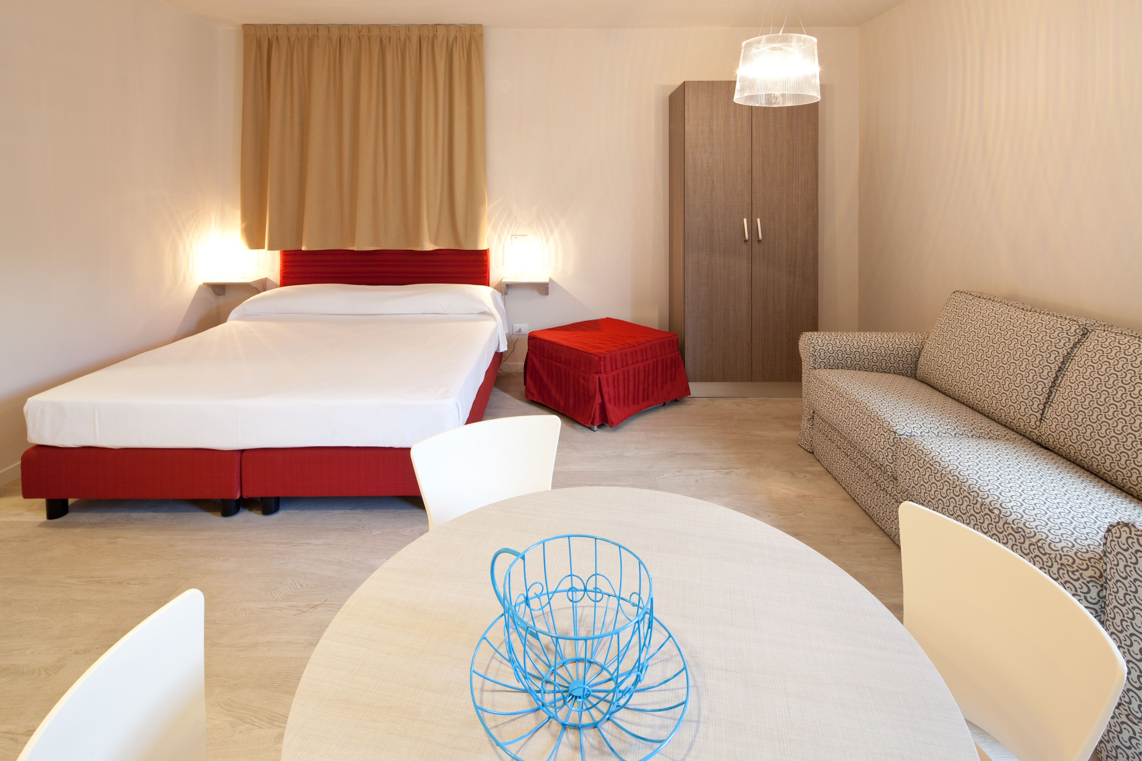 A hotel room with a bed, a red ottoman, a sofa, a round table with a blue wireframe object, and a wardrobe.