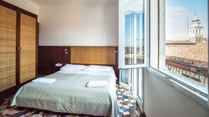 A hotel room with a bed, a wooden wardrobe, and a large window offering a view of a building with a dome. - Hotel Clodia (Chioggia)