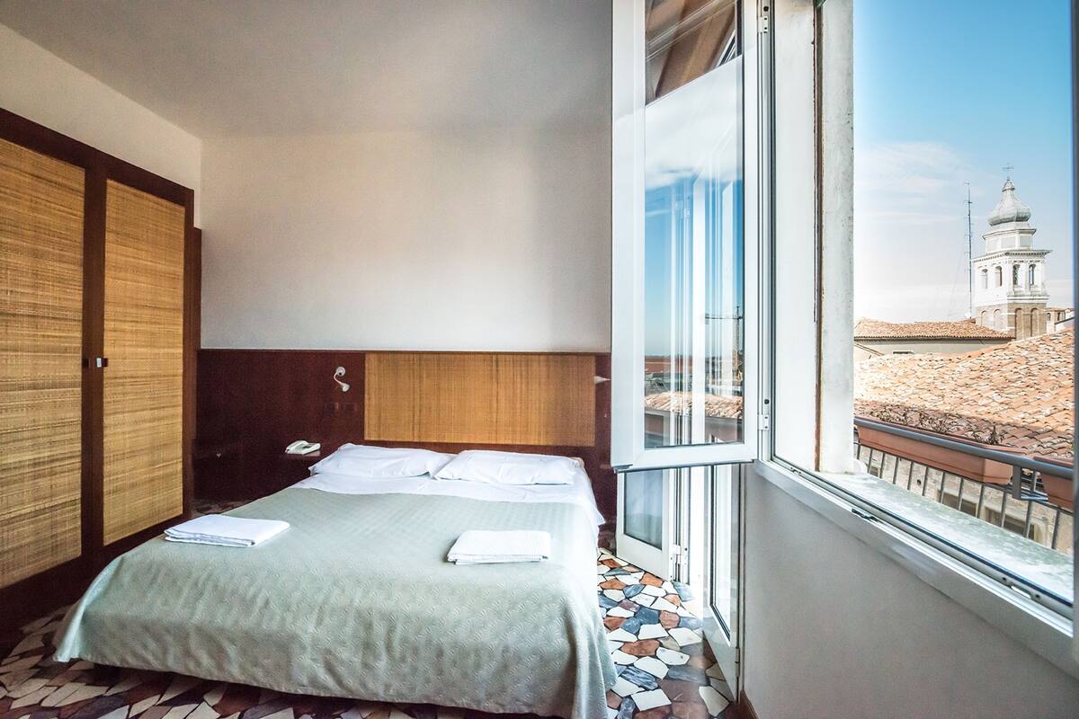 A hotel room with a bed, a wooden wardrobe, and a large window offering a view of a building with a dome.