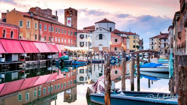 A canal with boats docked, buildings with red and white facades, and a tower with a clock. - Hotel Clodia (Chioggia)