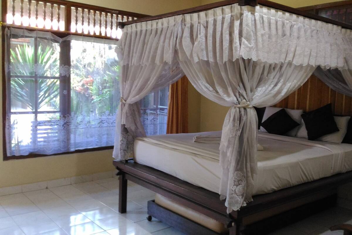 A four-poster canopy bed with a white bedspread and black pillows, positioned in a room with tiled flooring and a window with a view of greenery.