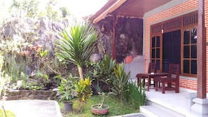 A garden with various plants and a brick building with a wooden porch and a table and chairs.
