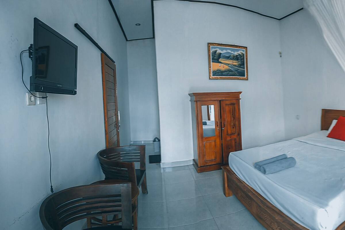A bedroom with a bed, a TV mounted on the wall, a wooden wardrobe, and a desk with a chair.