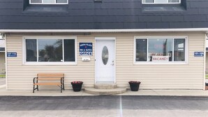A building with a beige exterior, a white door, and two windows. There are signs indicating 'Perth Plaza Inn & Suites Office' and 'Vacancy'. A bench and two flower pots are in front of the building.