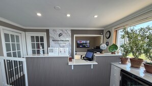 A reception desk with a computer, a TV displaying 'Ladies First,' a clock, and a sign for 'Perth Plaza Rooms & Suites.' - Perth Plaza Inn & Suites (Perth)