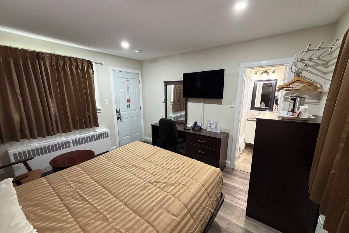 Une chambre d’hôtel comprenant un lit, un bureau avec un ordinateur, une télévision, un miroir, un porte-manteau et une fenêtre avec des rideaux.