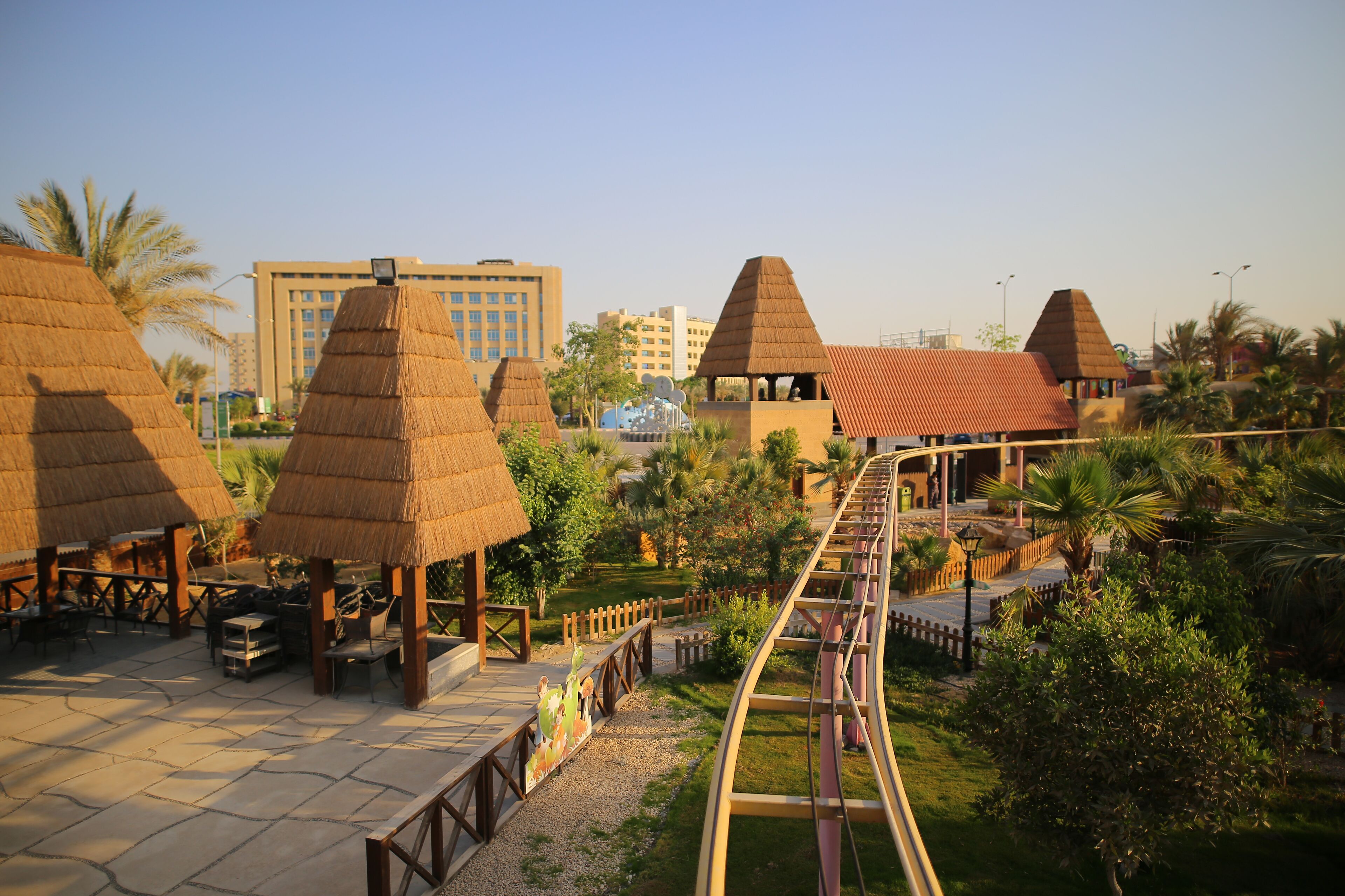 A theme park with thatched-roof structures, a roller coaster, and a paved walkway.