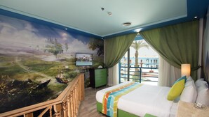 Habitación de hotel con un gran mural de un castillo en la pared, una cama con una manta colorida, cortinas verdes y vista al mar.