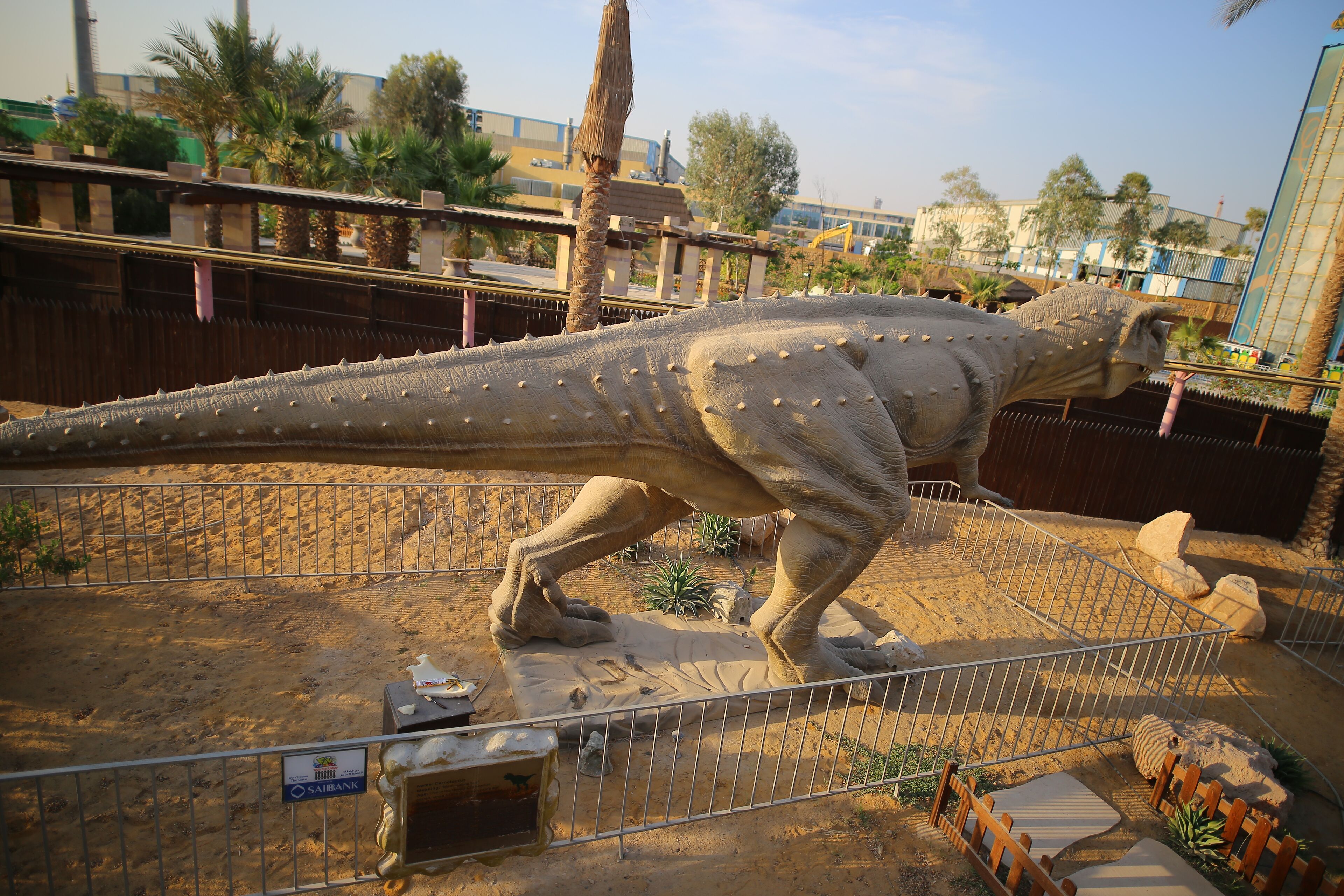 Una gran exhibición de dinosaurios con un recinto cercado, suelo arenoso y algo de vegetación.