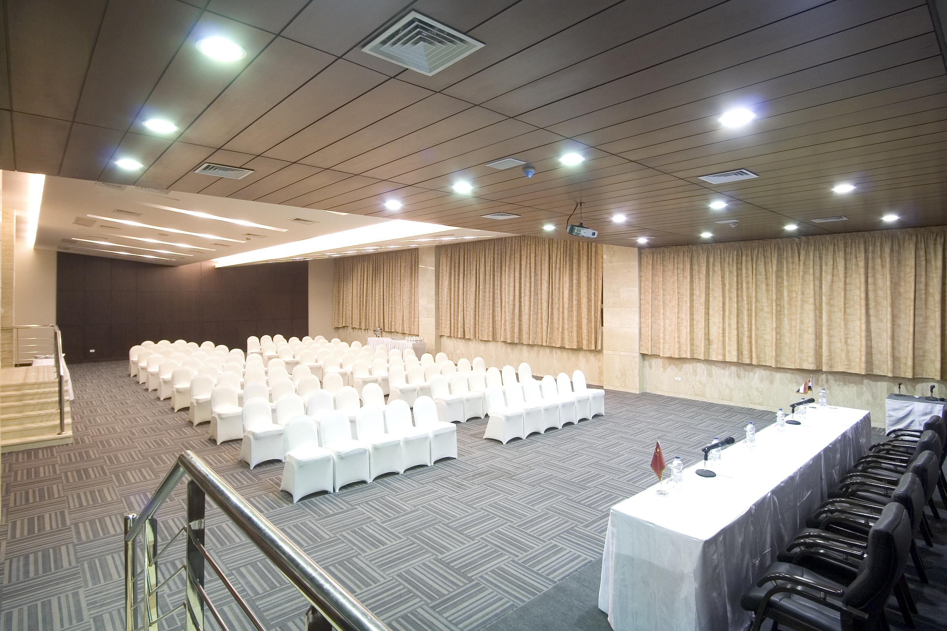 A conference room with rows of white chairs, a long table with microphones, and a projector screen.
