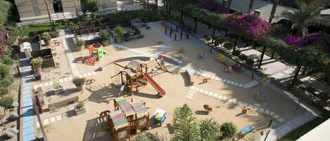 A playground with slides and swings, surrounded by palm trees and benches.