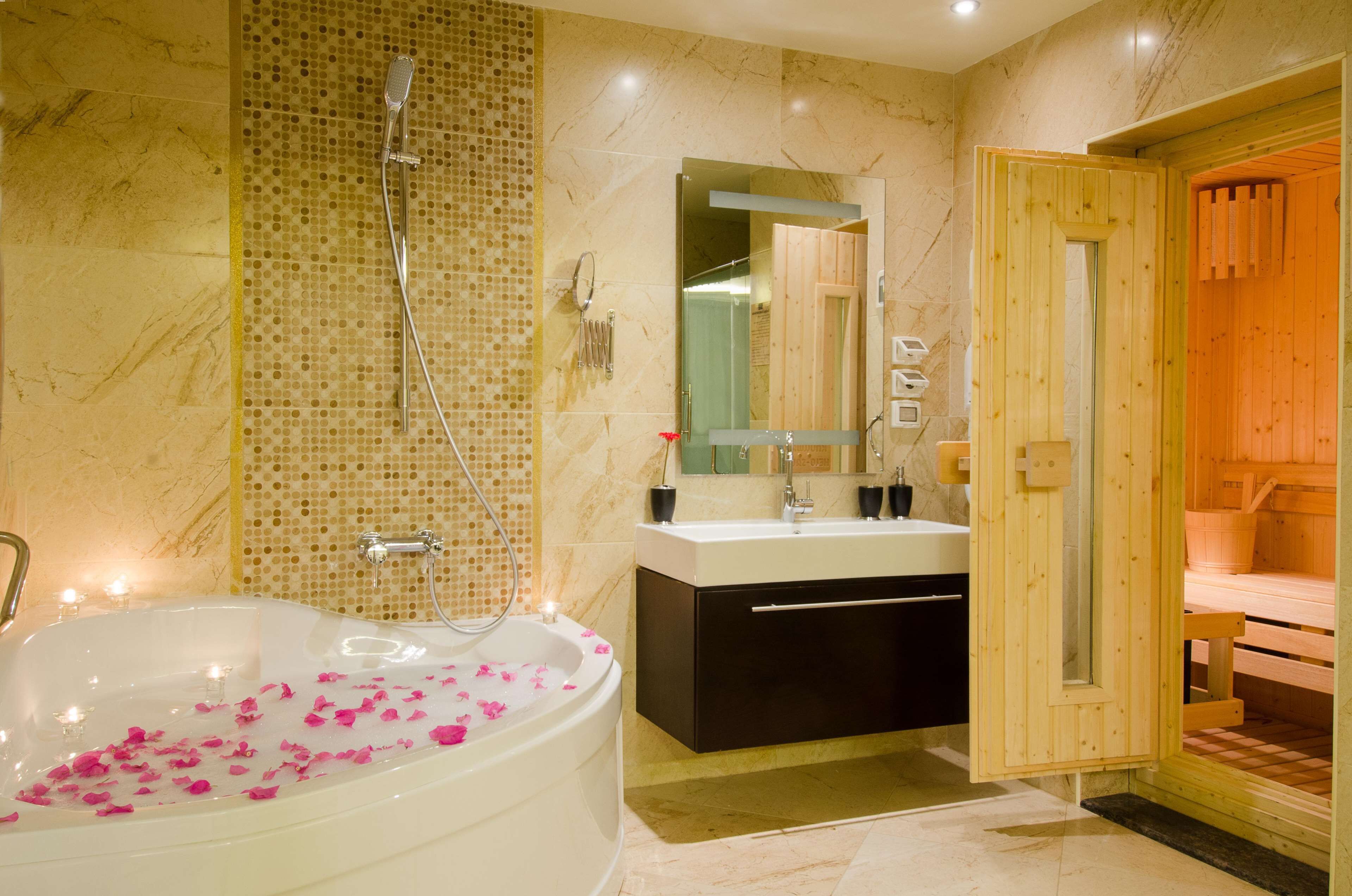 A modern bathroom with a large bathtub filled with rose petals, a wooden sauna, and a marble-tiled shower area.