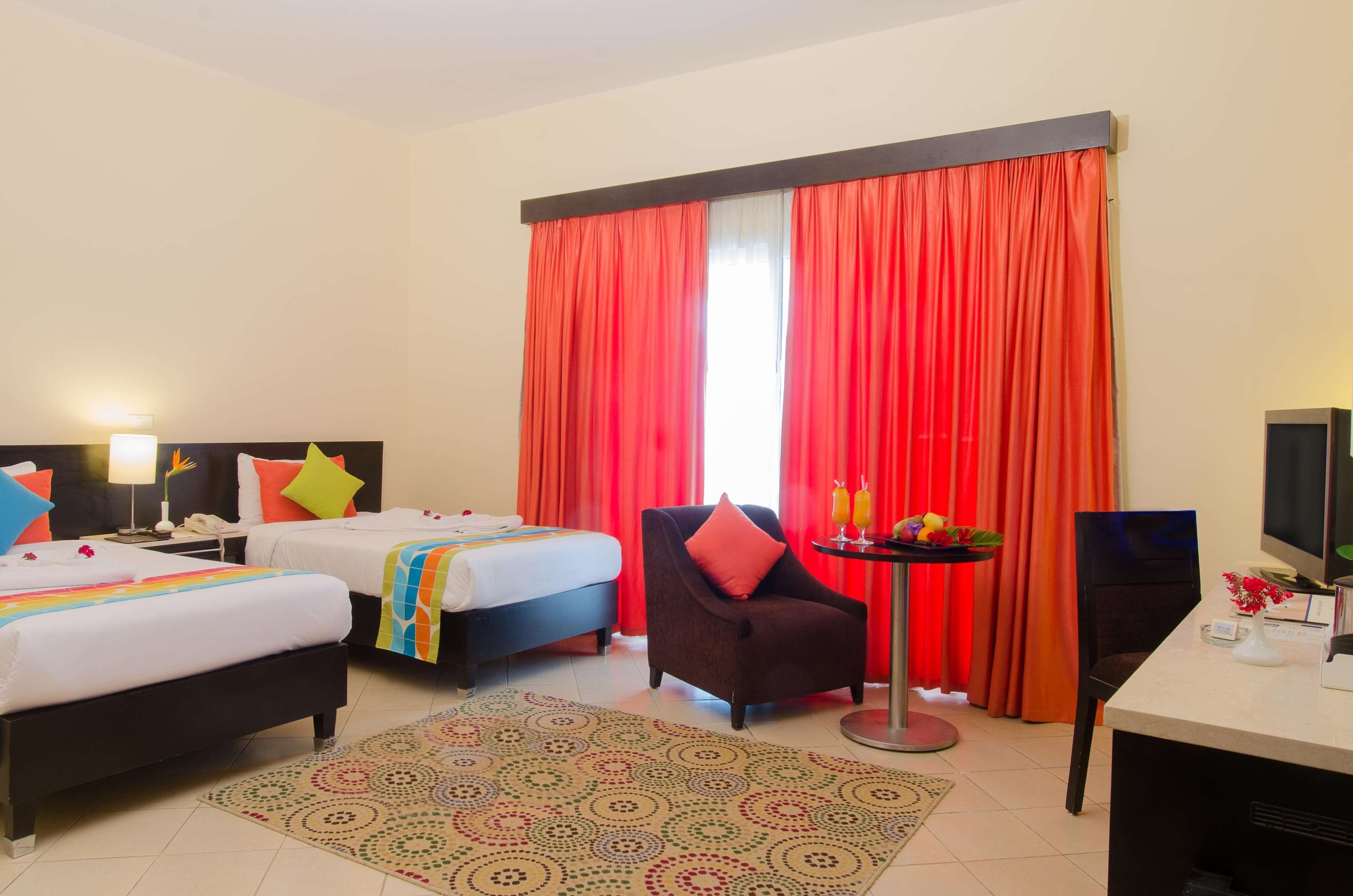 Habitación de hotel con dos camas, un escritorio, una silla, un televisor y una ventana con cortinas rojas.