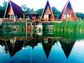 Three wooden triangular cabins with a wooden deck and a small bridge connecting them over a calm body of water.