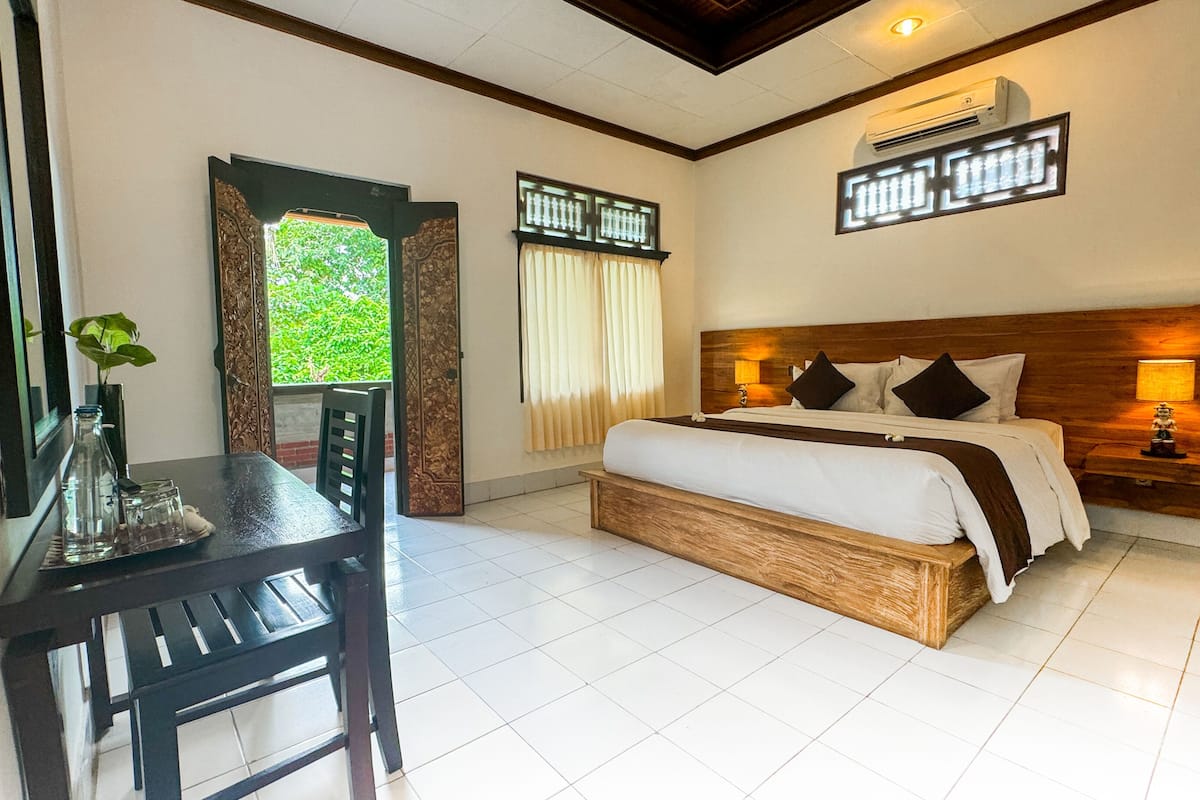 Quarto com cama de madeira, escrivaninha com um vaso e vista para o verde pela janela.