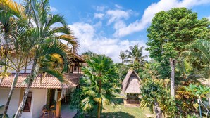 Una villa tropical con una cabaña de techo de paja, palmeras y un edificio blanco con tejado de baldosas.