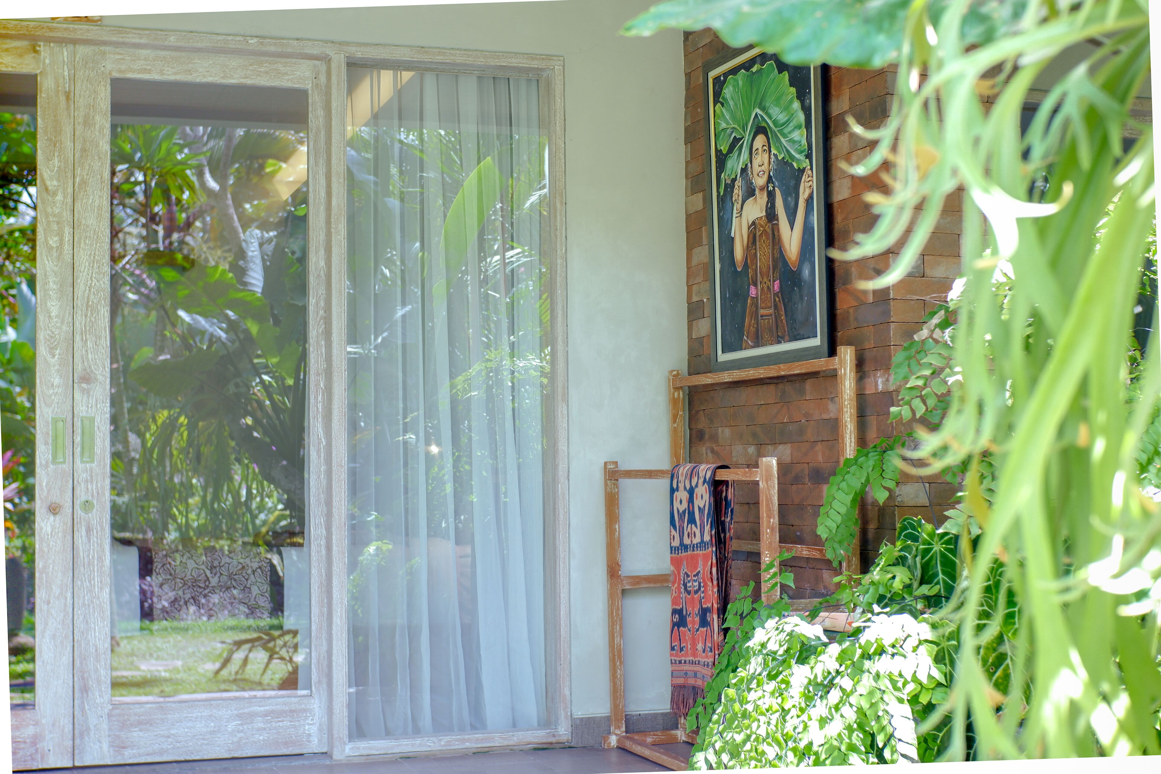 A room with a sliding glass door leading to a garden, a wooden bench, and a wall-mounted painting.