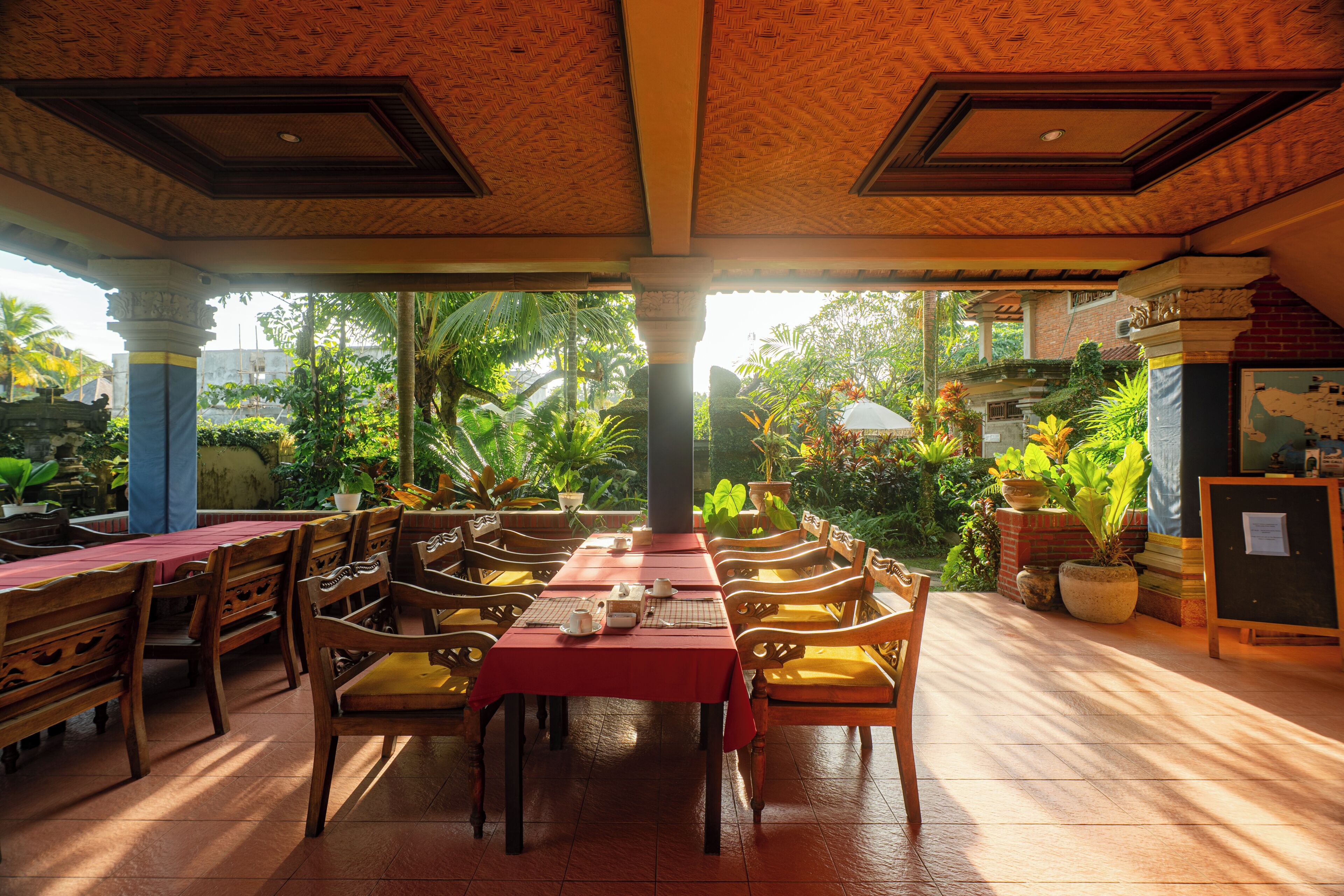 An indoor dining area with wooden tables and chairs, a red tablecloth, and potted plants.