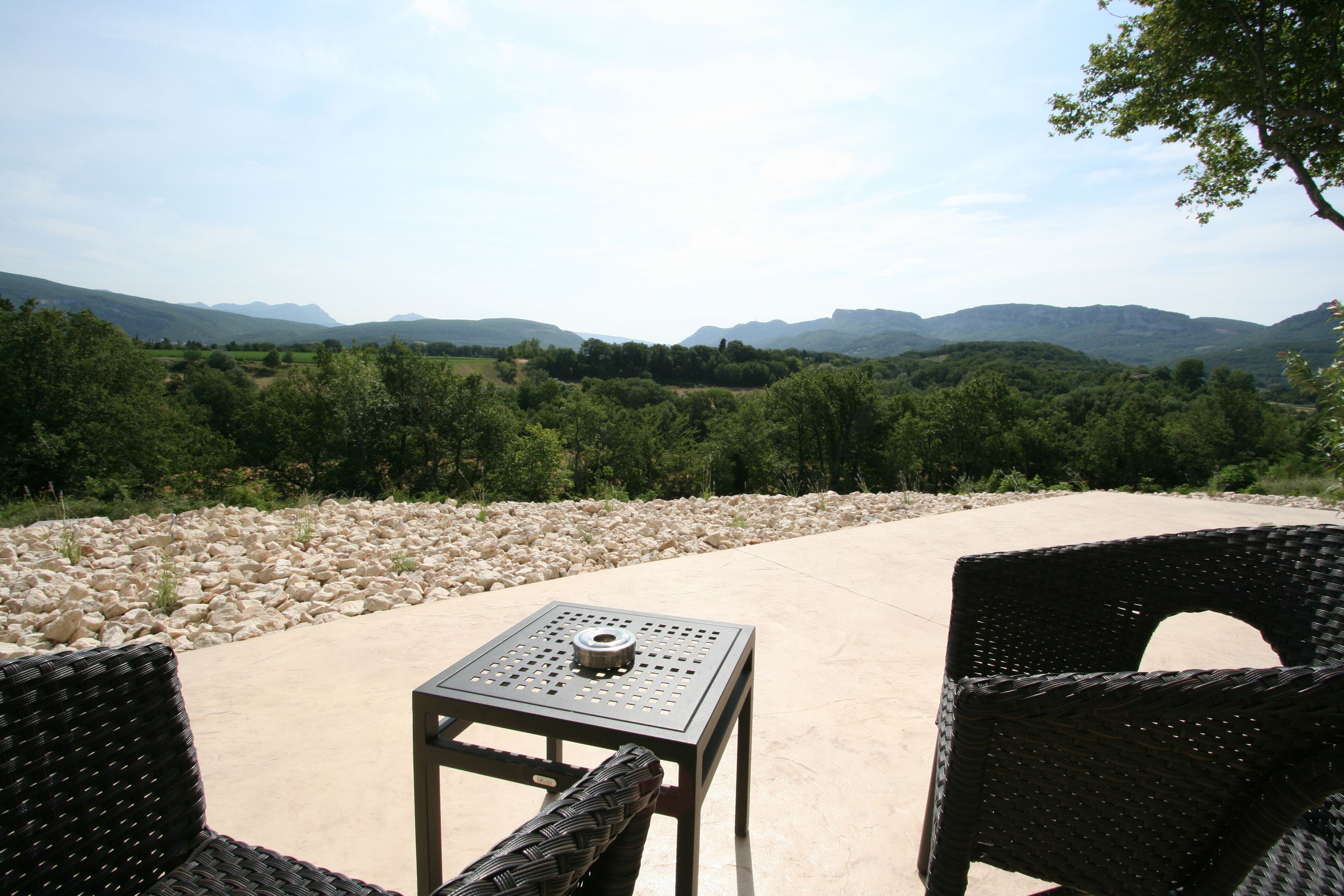 An outdoor seating area with wicker chairs and a metal table, overlooking a mountainous landscape.