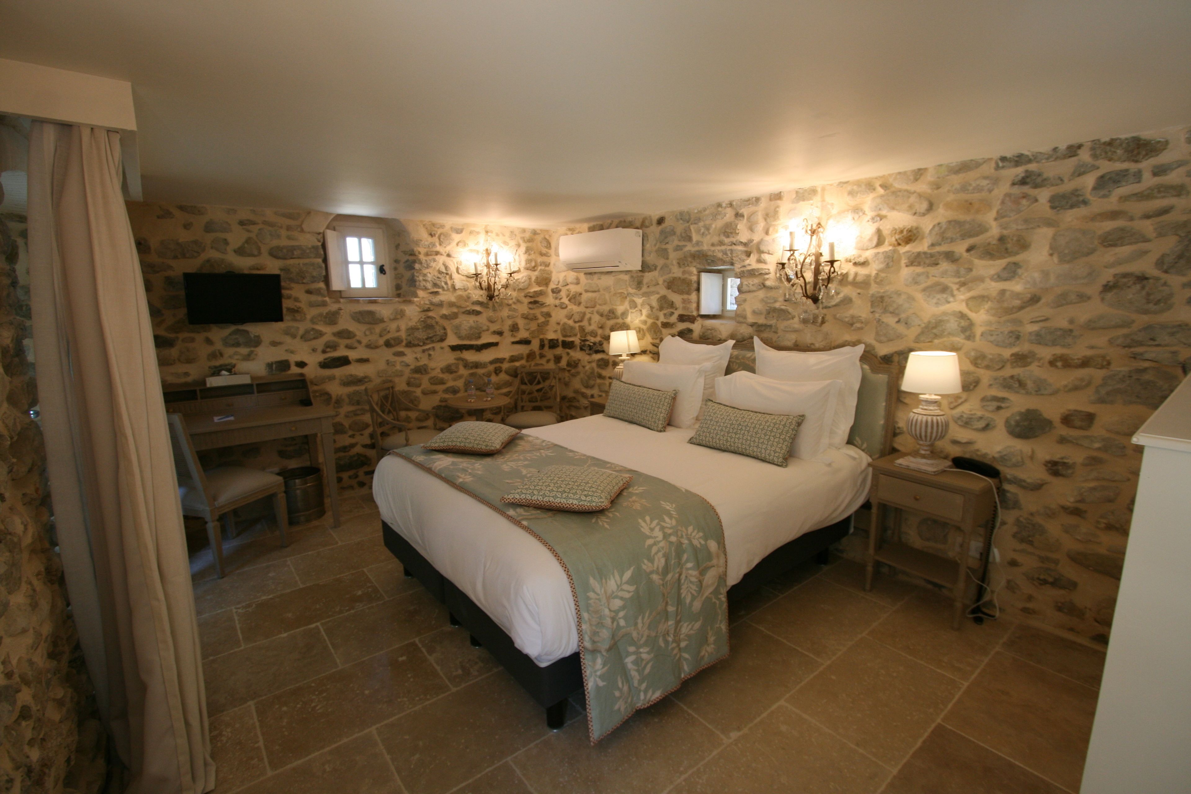 A bedroom with a stone wall, a bed with a green bedspread, bedside lamps, a desk, and a television.