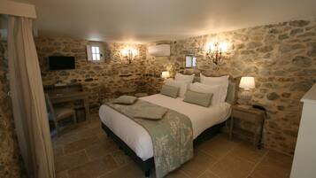 A bedroom with a stone wall, a bed with a green bedspread, bedside lamps, a desk, and a television.