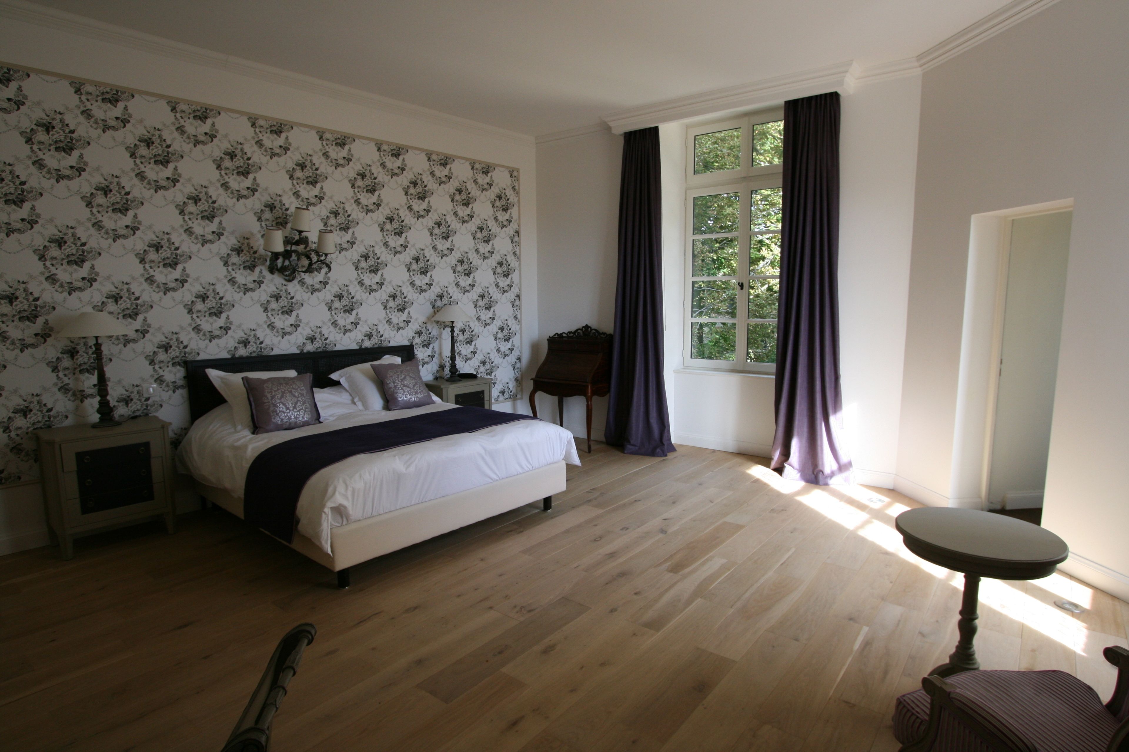 Ein Schlafzimmer mit einem großen Bett, einem Nachttisch, einem Sessel, einem kleinen Tisch und einem Fenster mit Vorhängen.