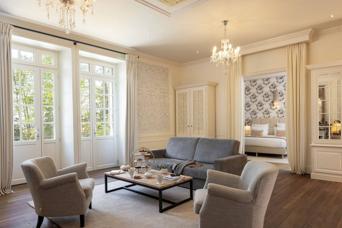 A spacious living room with a grey sofa, two armchairs, a coffee table with decorative items, and a chandelier.