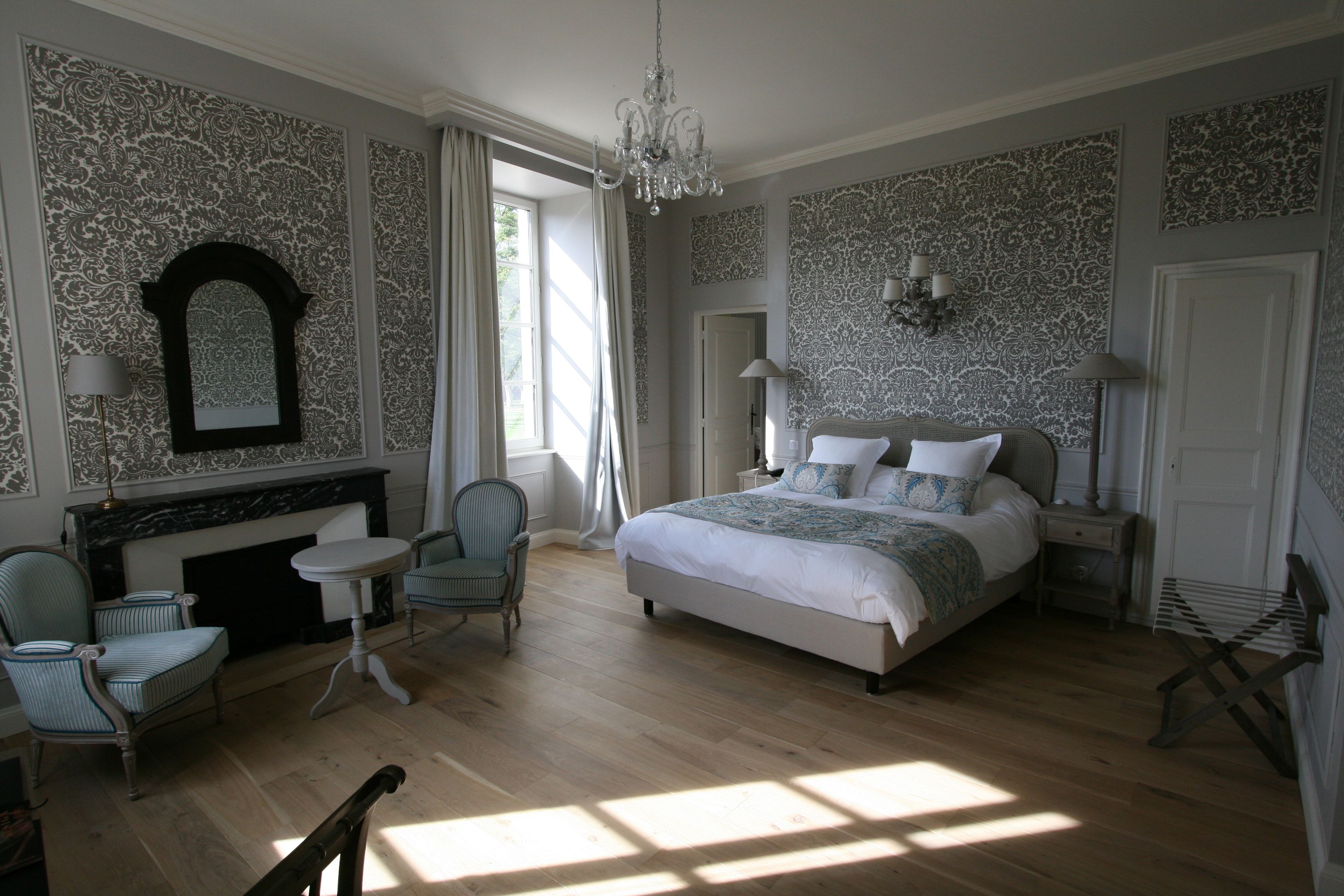 A bedroom with a large bed, a fireplace, a chandelier, and patterned wallpaper.
