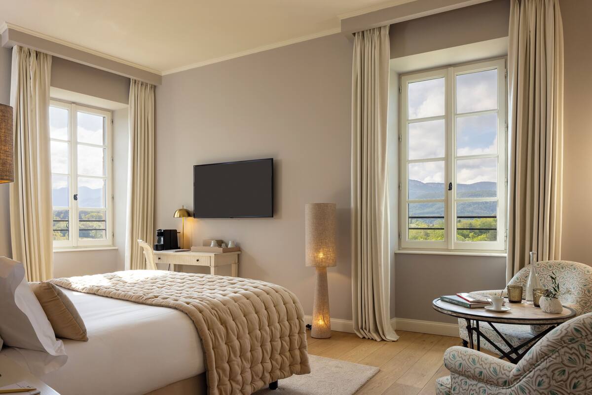 A hotel room with a bed, a desk with a lamp, a chair, a table with a vase, and a TV.