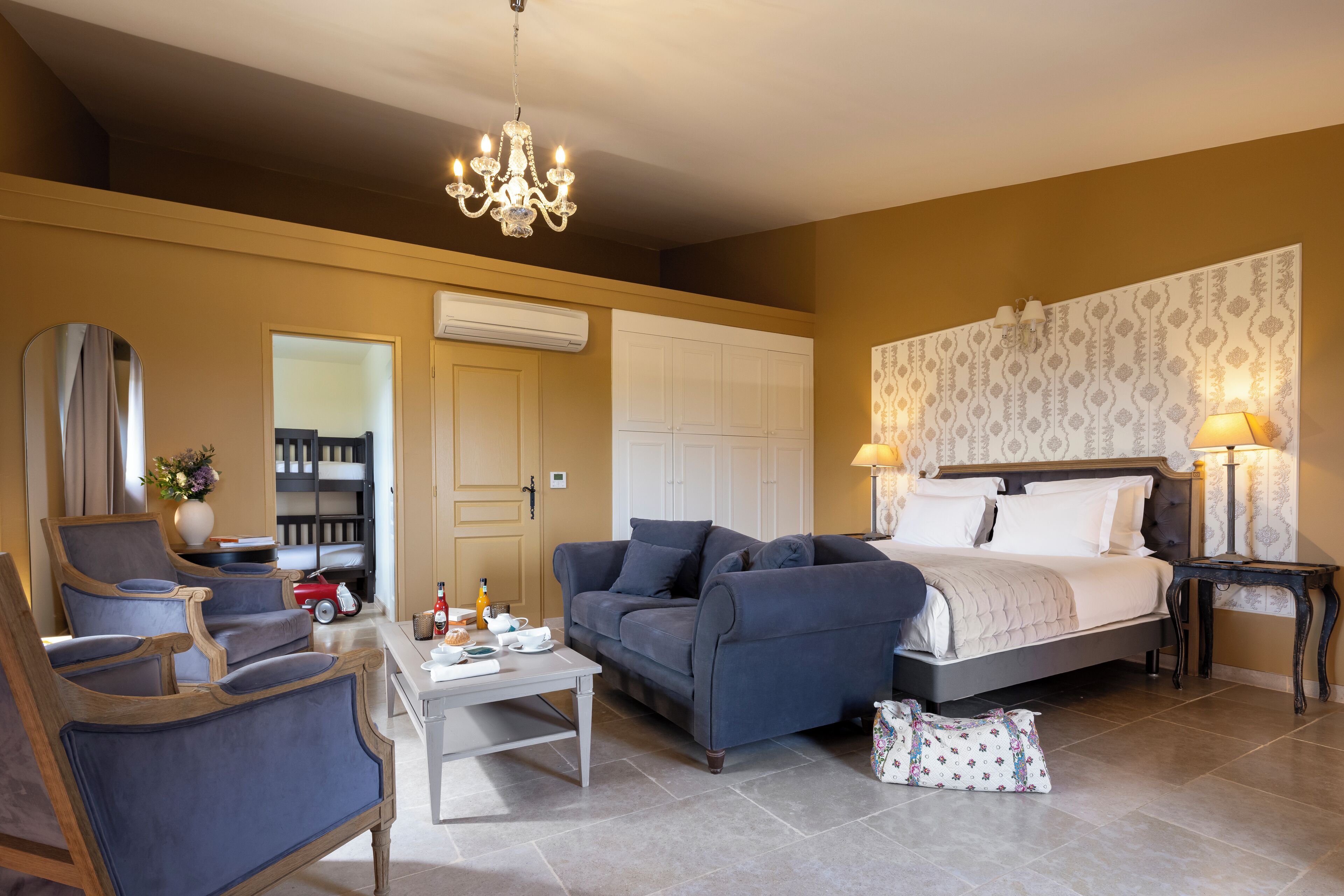 A room with a bed, a sofa, a coffee table, and a chair. There is a chandelier hanging from the ceiling.