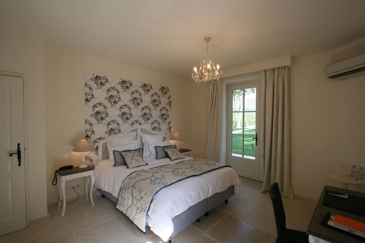 A bedroom with a large bed, a nightstand, a chair, a desk, a chandelier, and a window with curtains.