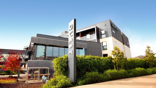 A modern hotel with a prominent 'QUEST' sign, large windows, and a well-maintained garden area.
