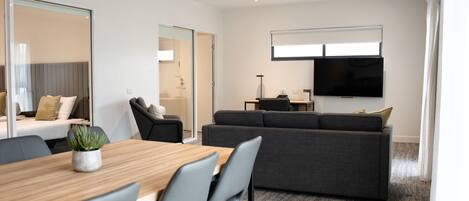 A modern hotel room with a wooden dining table, grey chairs, a grey sofa, a flat-screen TV mounted on the wall, and a glass partition separating the living area from the bedroom.