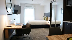 A modern hotel room with a wooden desk, a laptop, a chair, a bed with pillows, a TV, and a large mirror.