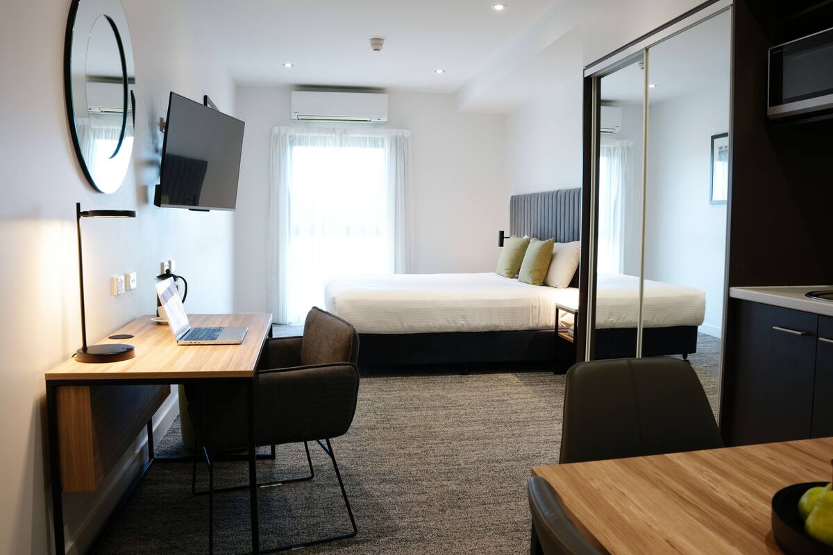 A modern hotel room with a wooden desk, a laptop, a chair, a bed with pillows, a TV, and a large mirror.
