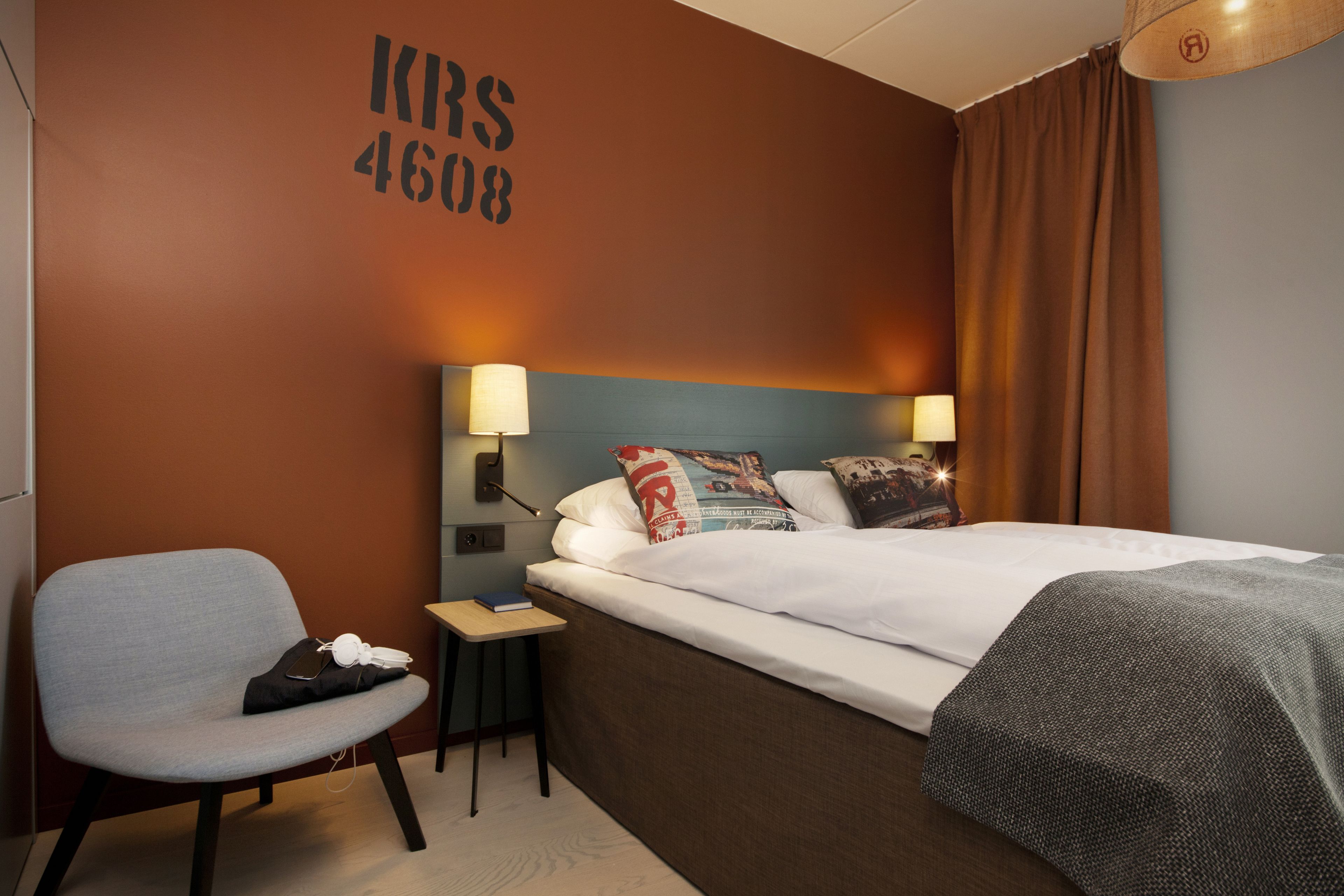 A hotel room with a bed, a chair, a nightstand, and a wall with a number.