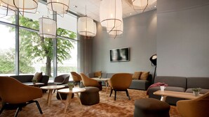 Ein moderner Lounge-Bereich mit Sofas, Sesseln und Tischen, große Fenster mit Blick auf Bäume und ein an der Wand montierter Flachbildfernseher.