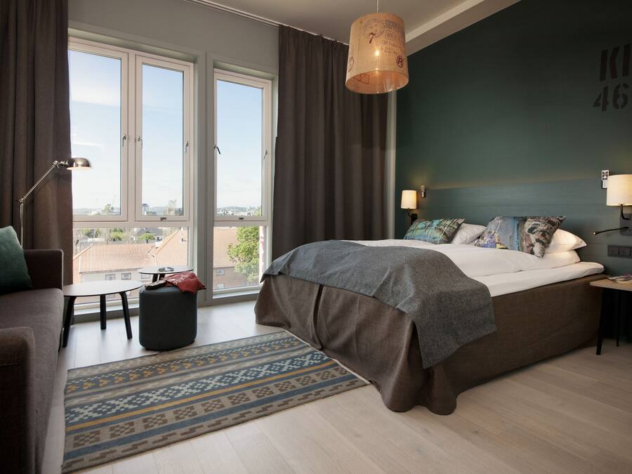 A bedroom with a large bed, a sofa, a small table, a lamp, and a view of the city through the window.