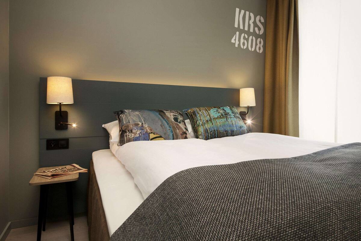 Une chambre d’hôtel avec un grand lit, deux lampes de chevet, une table de chevet avec un journal et un mur portant le numéro « 4608 ».