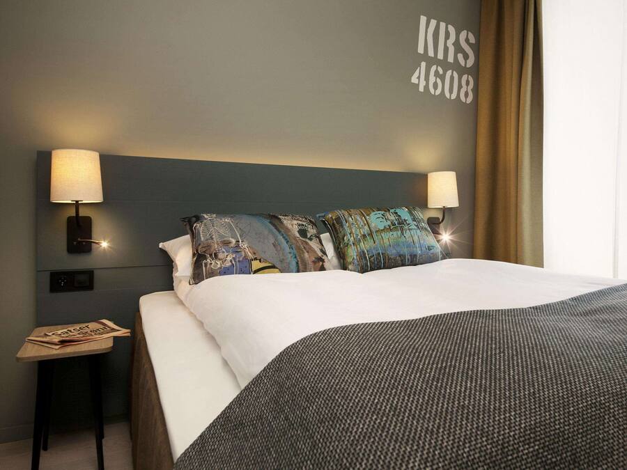 A hotel room with a large bed, two bedside lamps, a nightstand with a newspaper, and a wall with the number "4608".