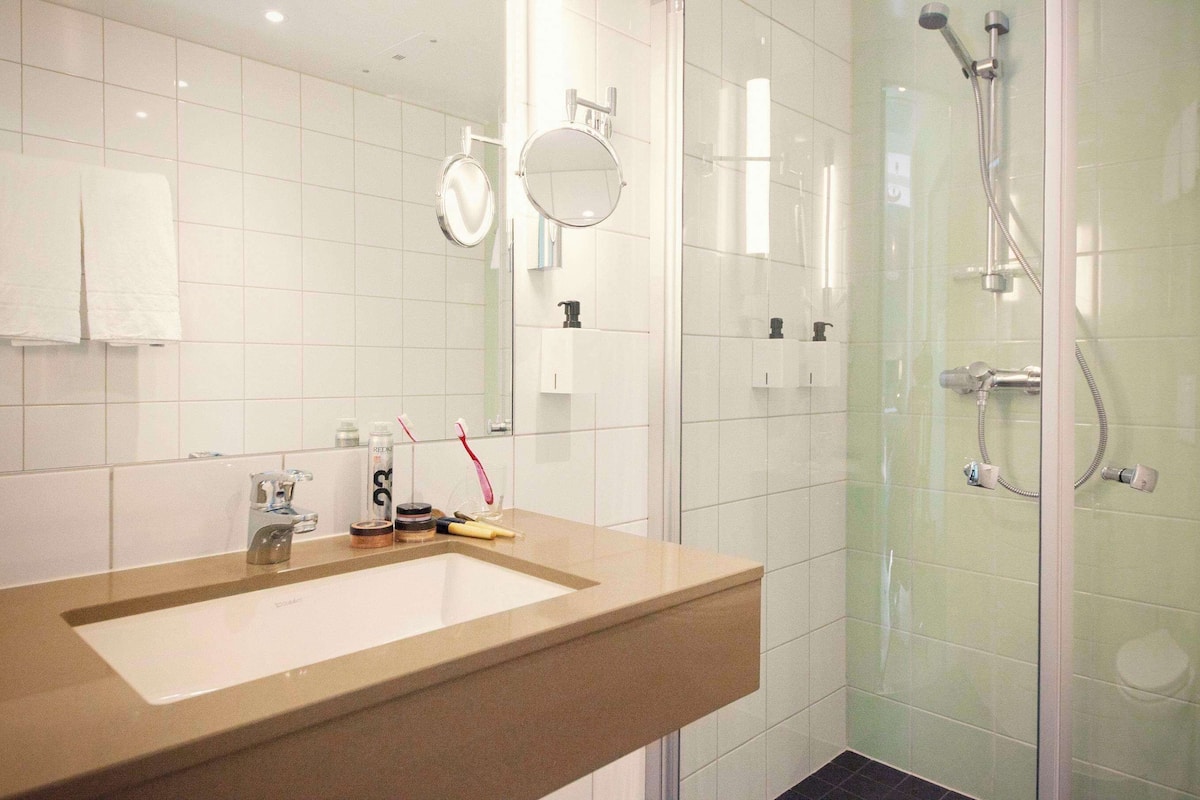 Une salle de bain avec un lavabo beige, deux miroirs, une cabine de douche en verre et une serviette blanche accrochée au mur.