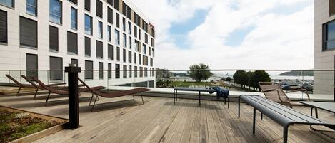A rooftop terrace with wooden flooring, sun loungers, and a view of a modern building and a body of water.