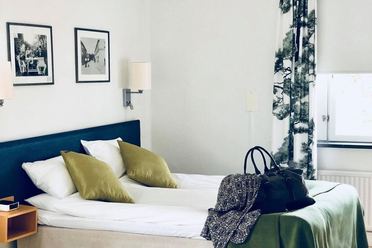 A neatly arranged bedroom with a bed, pillows, a bedside table, a lamp, a chair, a rug, and a window with curtains.