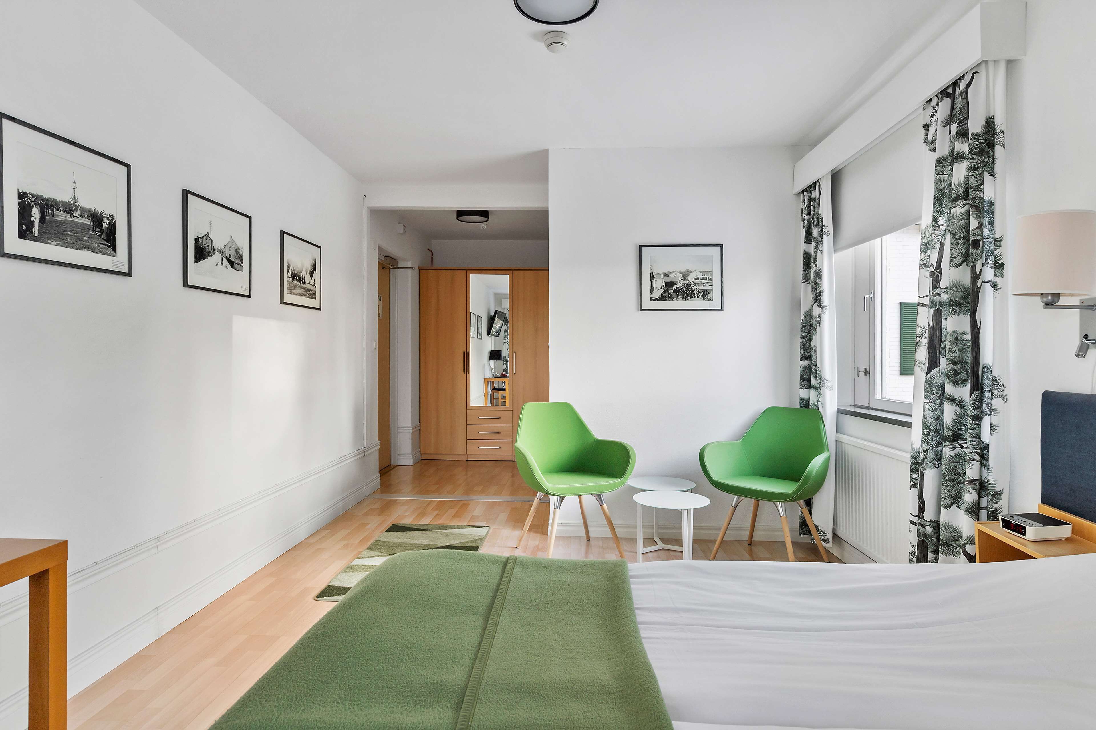 Habitación de hotel moderna con una cama, sillas verdes, una mesita y cuadros en las paredes.