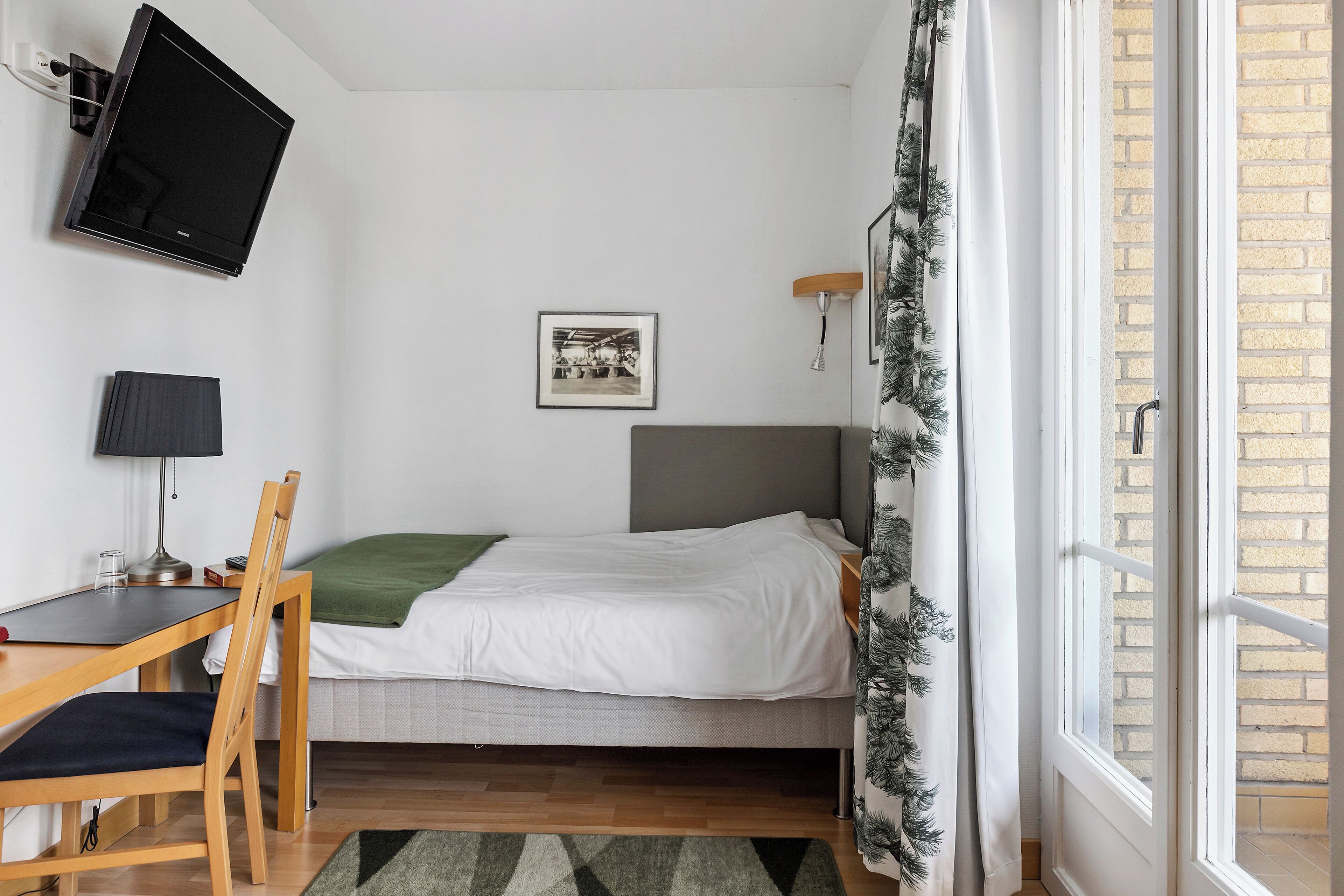 A bedroom with a bed, a desk with a chair, a TV mounted on the wall, and a window with curtains.