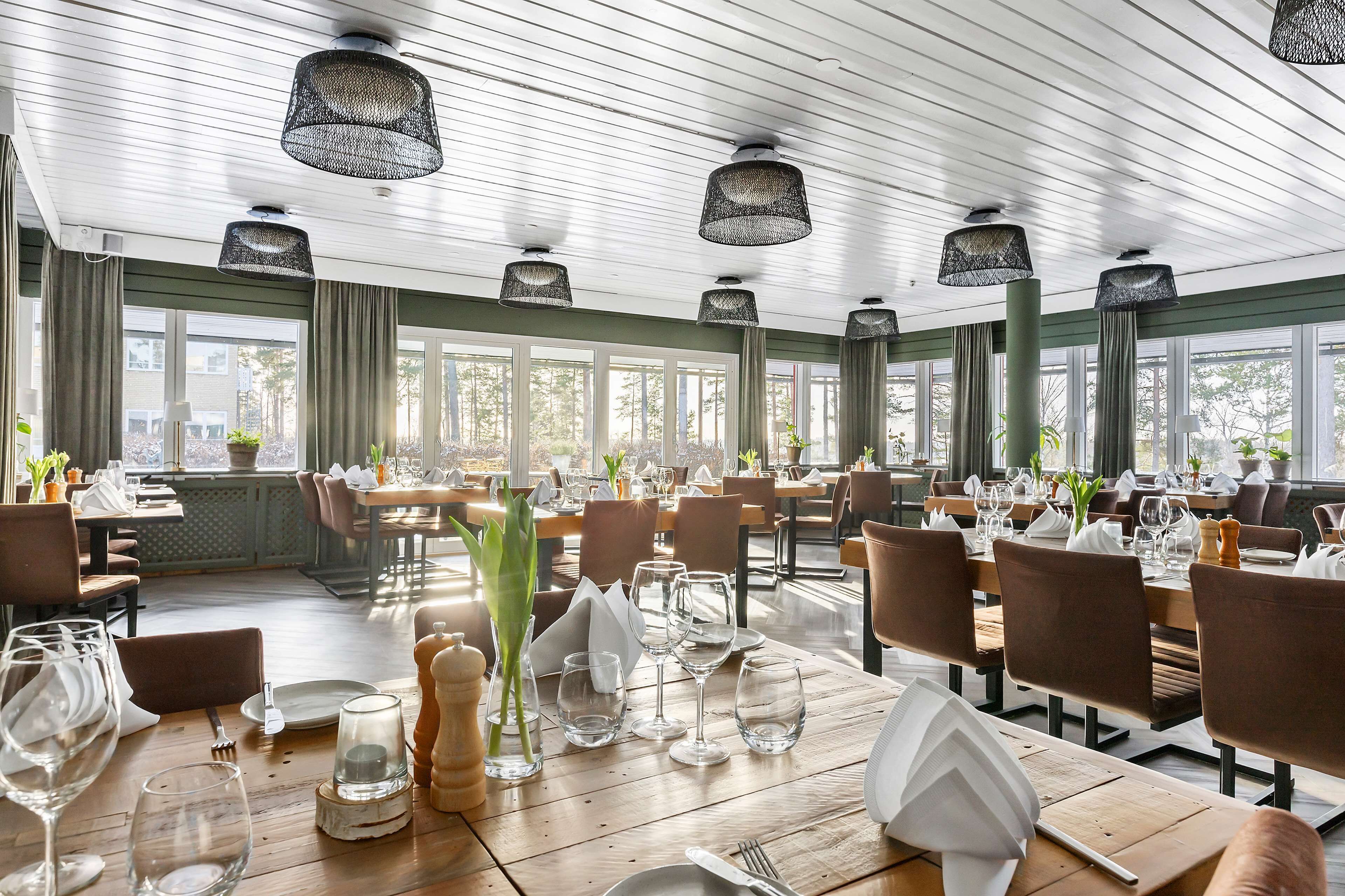 Een restaurant met houten tafels, zwarte hanglampen en grote ramen.