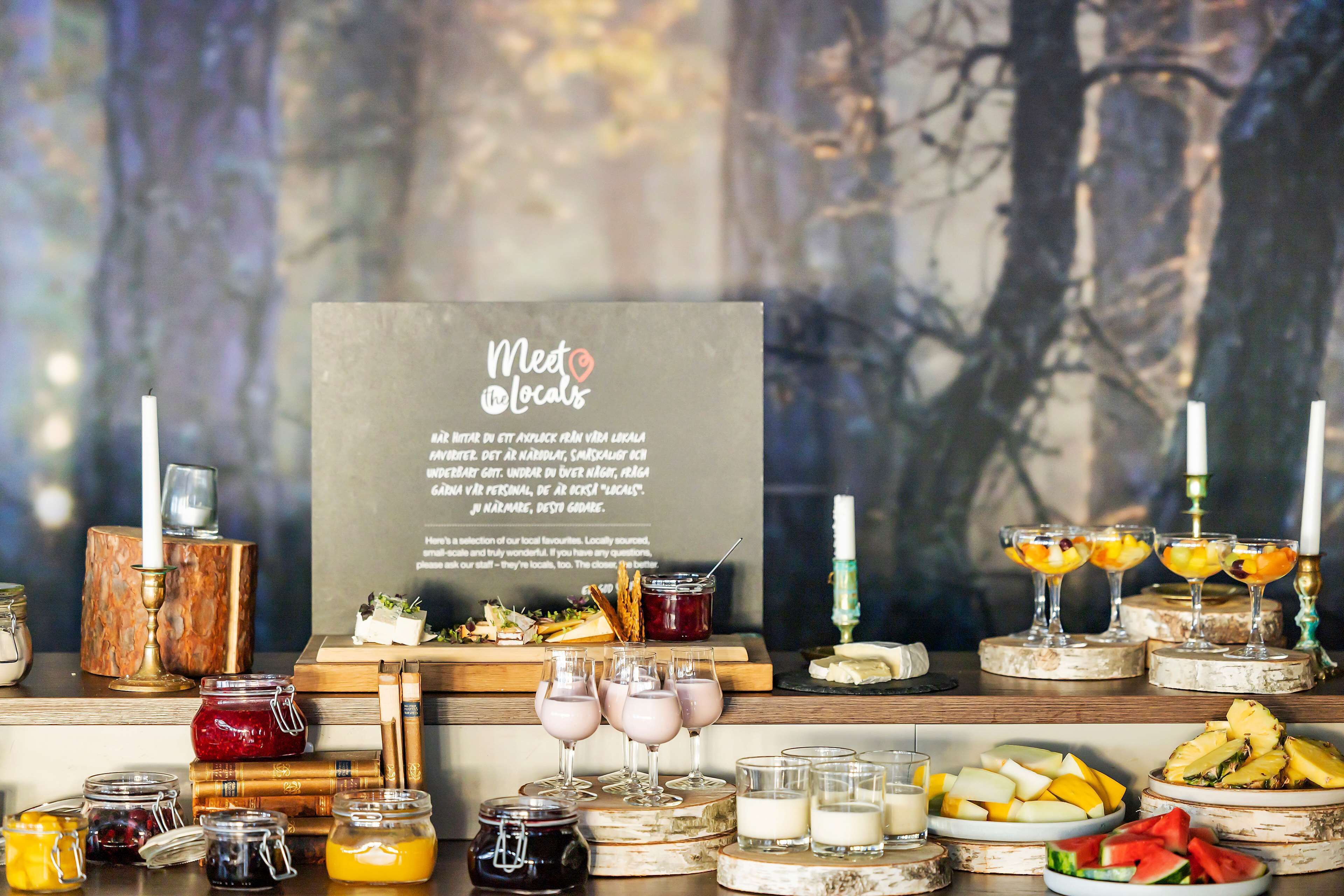 A buffet setup with various food items and drinks, including jars of preserves, a selection of fruits, and glasses of beverages.